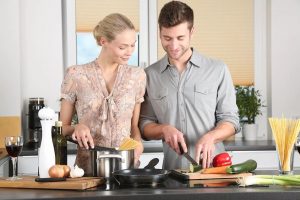 A man and woman cooking together in a kitchen with food and cooking supplies around them on the stove and counter.