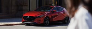 A red 2020 Mazda3 Hatchback parked in front of a building with a woman's image blurred to the right, up close to the lens. | Mazda dealer in St. Louis, MO