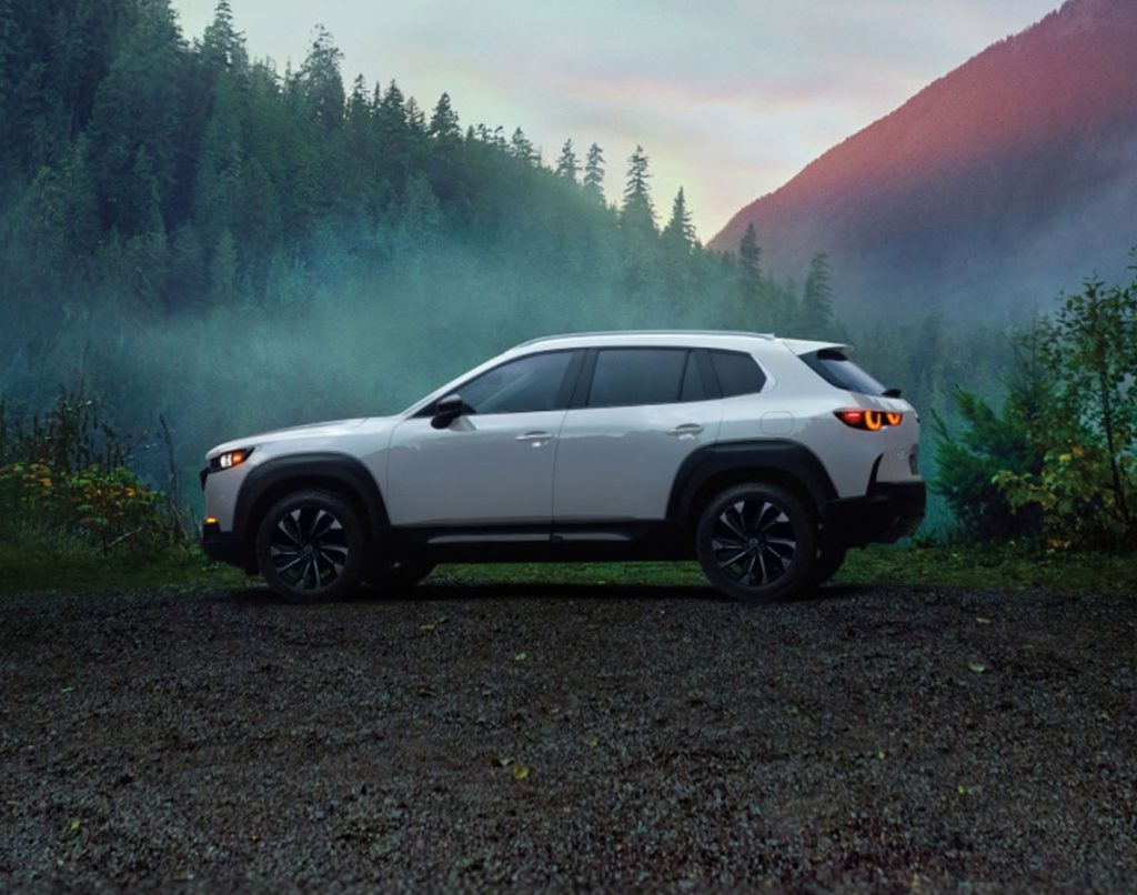 White Mazda CX-50 parked on a gravel area with forested mountains in the background.