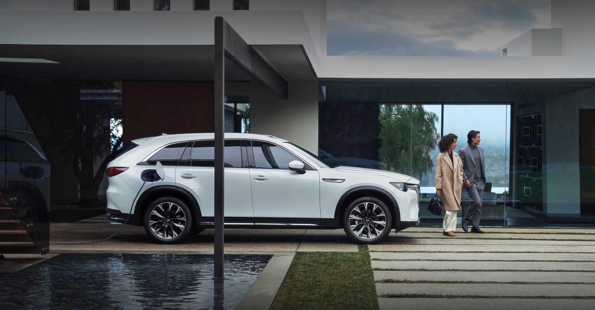 White Mazda CX-90 SUV parked outside a modern home with people nearby.